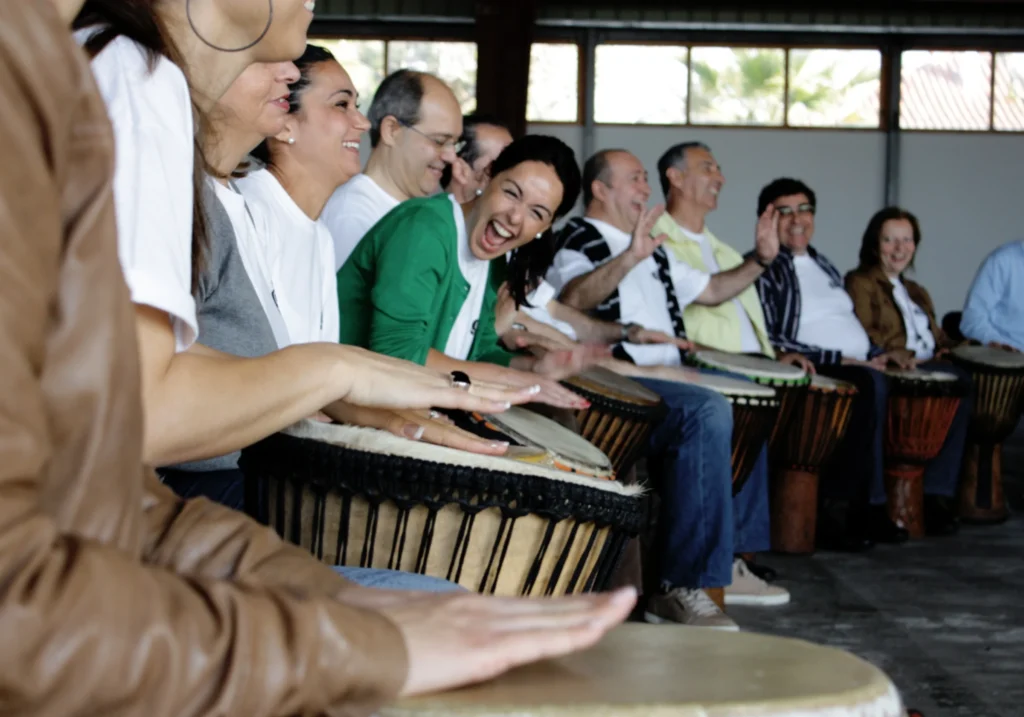 Group of people playing djembe drums and laughing during a percussion team building session