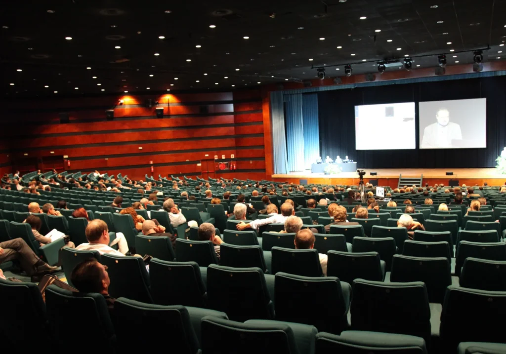 Large auditorium full of attendees during a conference with speakers on stage and screens