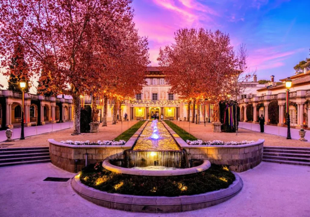 Mansion exterior with illuminated fountain and gardens under a pink and purple sunset sky