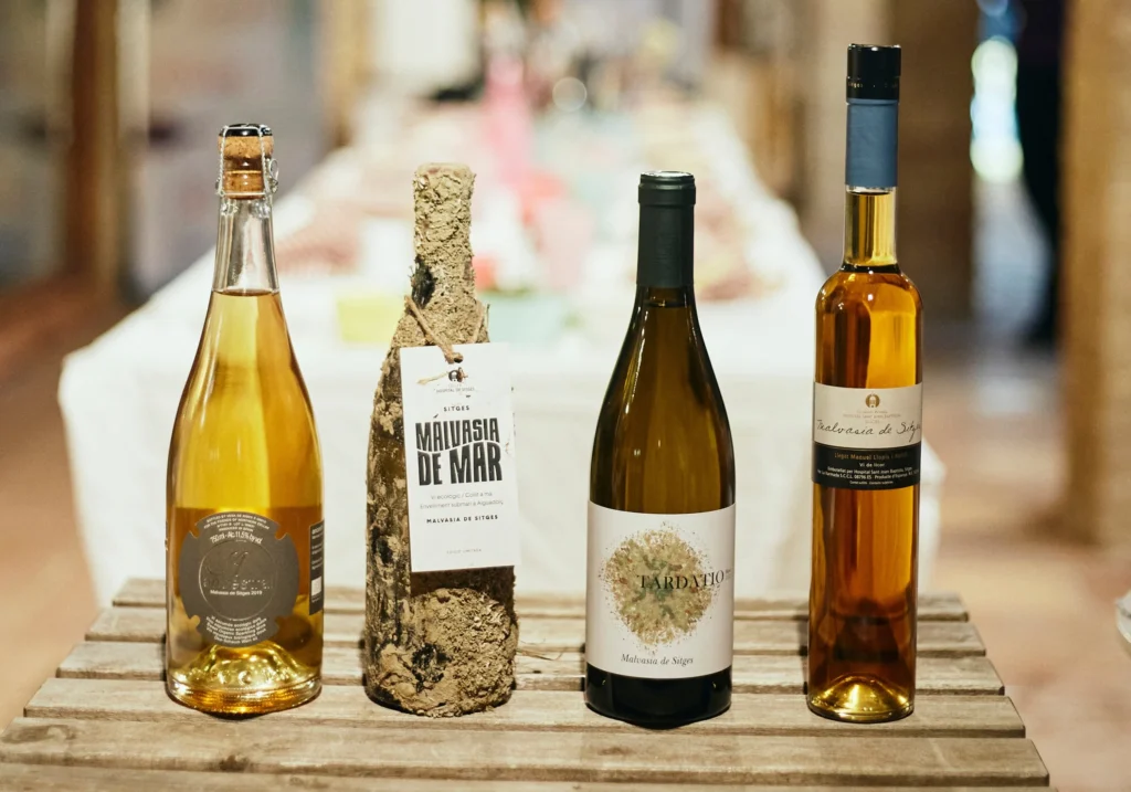 Four bottles of Sitges Malvasia wine, including the iconic Malvasia de Mar, on a wooden surface